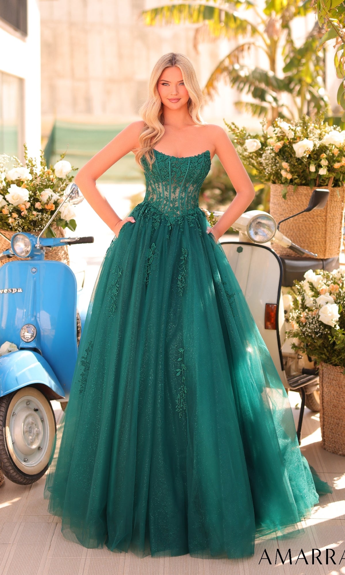 A woman stands outdoors in the Strapless Long Prom Ball Gown, Amarra 88913, a green dress with a sheer lace corset, near a blue scooter and flower arrangements.